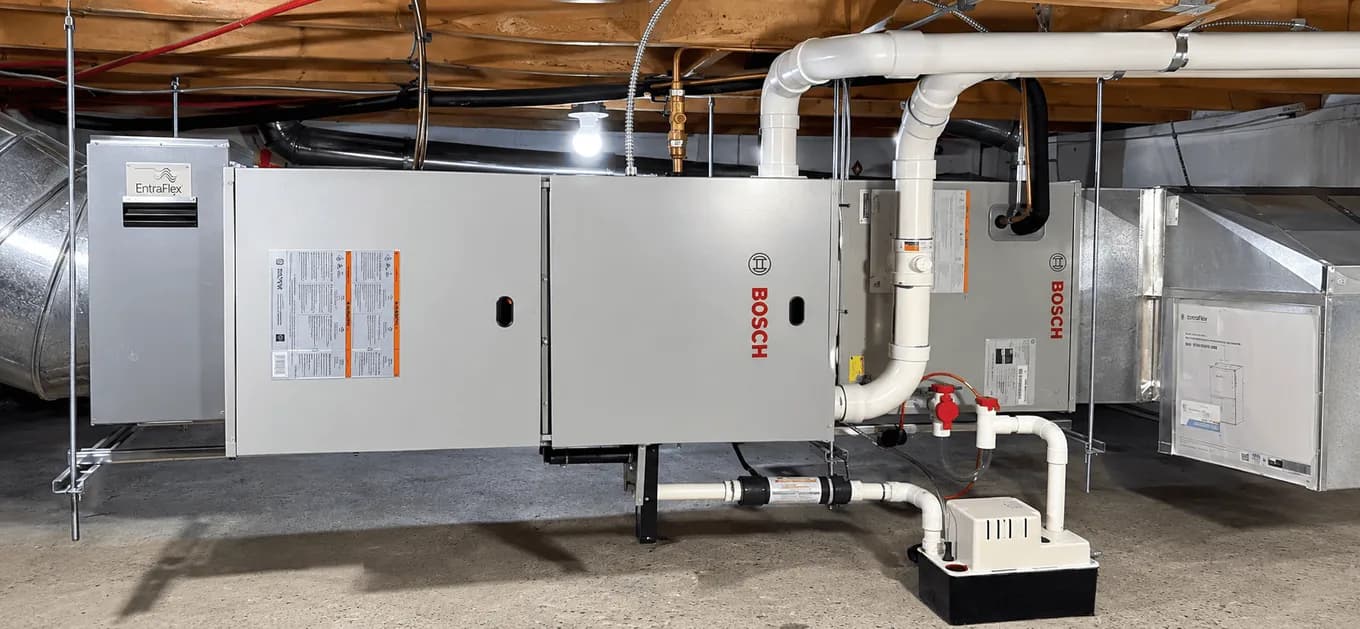 Two indoor air handlers installed side by side in a basement, tied into galvanized supply ducting, with a condensate pump on the floor and a ventilator mounted on the wall alongside