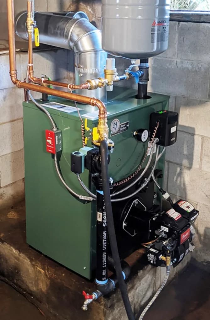 A green cast iron oil fired boiler in a basement, with copper supply piping run across the top, an expansion tank hung overhead and a galvanized flue pipe turned into the chimney